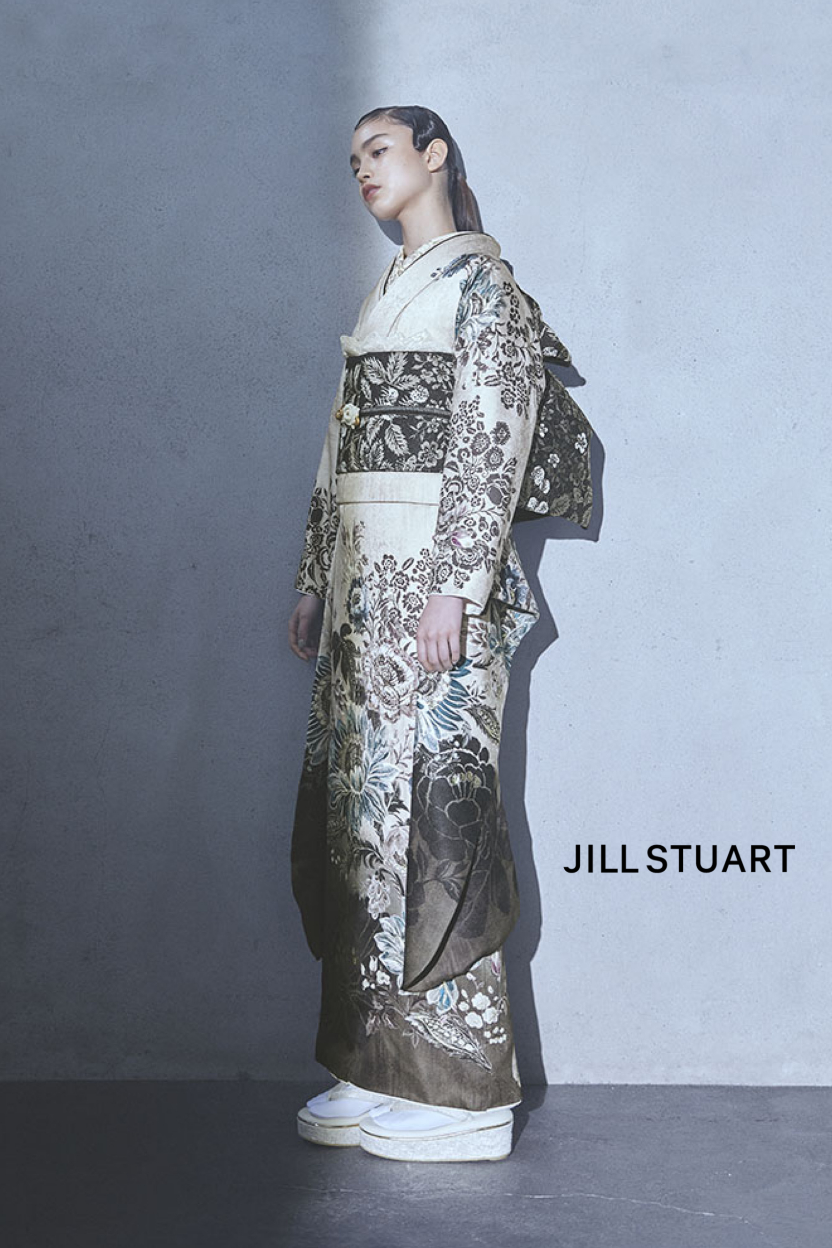 灰色の壁の横に「JILL STUART」と商品名「JI112」がさりげなく刻まれている人物が立っており、緑、灰色、白の色合いで複雑なデザインが施された花柄の着物を着て、後ろに大きなリボンが付いた濃い色の帯と白い厚底靴を合わせている。