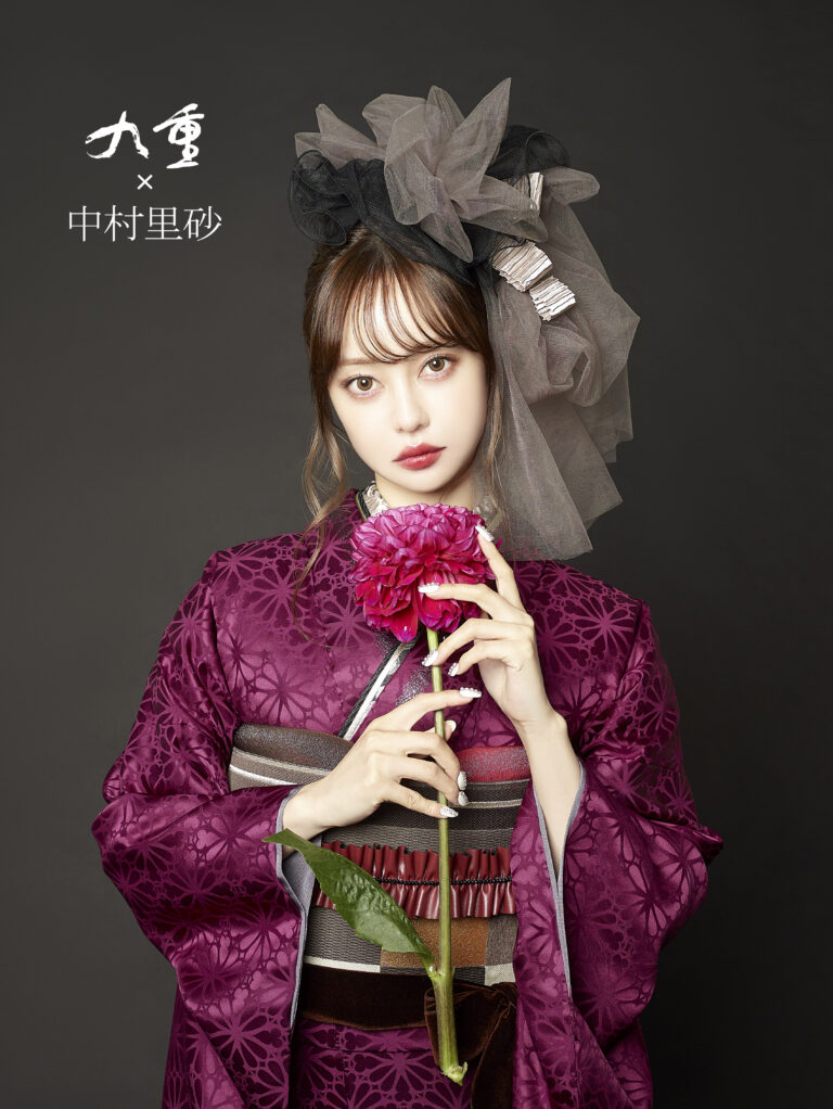 手の込んだ紫色の花柄の着物を着た女性が、ピンクのダリアを唇に当てています。彼女は、銀色のアクセントで飾られた大きな黒とグレーのチュールのヘッドピースをかぶっています。背景は暗い無地で、左側に日本語の文字があります。