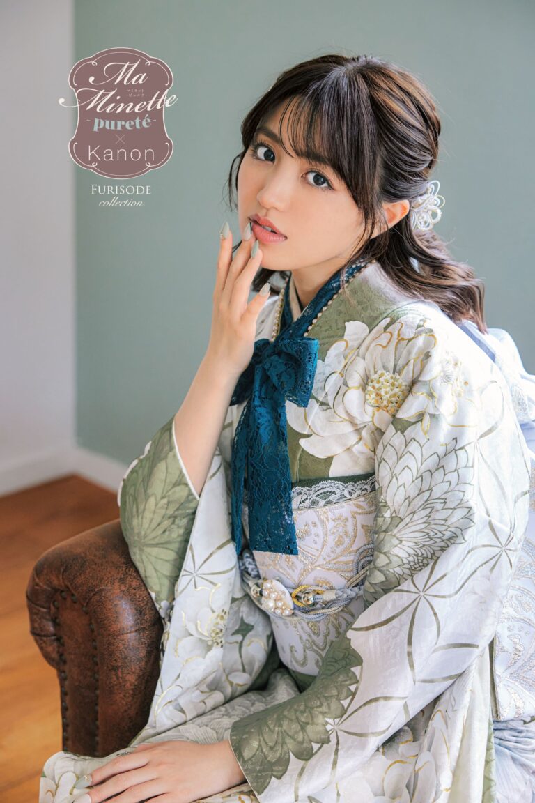 緑と白の複雑な花柄が描かれた上品な振袖を着た女性が室内に座っている。長い黒髪を飾りクリップでまとめ、首には青いレースのリボンをつけている。顔の近くに手を置き、カメラを優しく見つめている。背景には落ち着いた緑の壁がある。