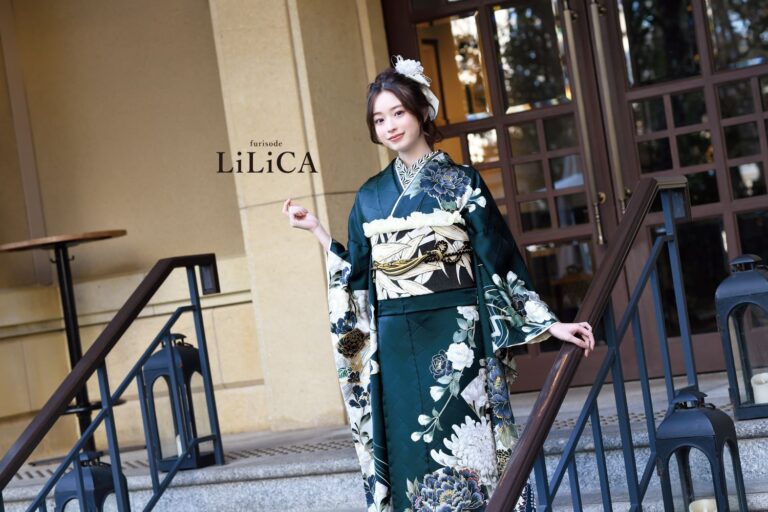 濃い緑色の花柄の着物に淡い色の帯を締め、小さなアクセサリーを持った人物が屋外の階段に立っています。ベージュ色の壁、大きな窓、暗い色の手すりなど、雰囲気はエレガントです。花柄のヘアピースをしており、近くに「LiLiCA」という文字が見えます。.