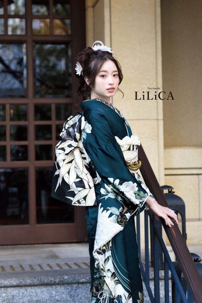 白い花柄の濃い緑の着物を着た女性が優雅に立っています。彼女の髪は白い花のアクセサリーでエレガントにスタイリングされています。彼女の後ろには木製のドアとベージュの壁がある建物があります。画像では、彼女の近くに「LiLiCA」という文字が見えます。.