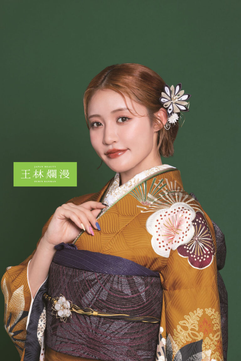 濃い緑色の背景に、花柄の華やかなマスタードイエローの着物を着た女性が立っています。彼女の髪は白と紫の花のヘアピースできれいにスタイリングされています。彼女は片手を胸の近くで上げ、優しく微笑んでいます。彼女の横には「ミス キモノ 2023」という文字があります。.