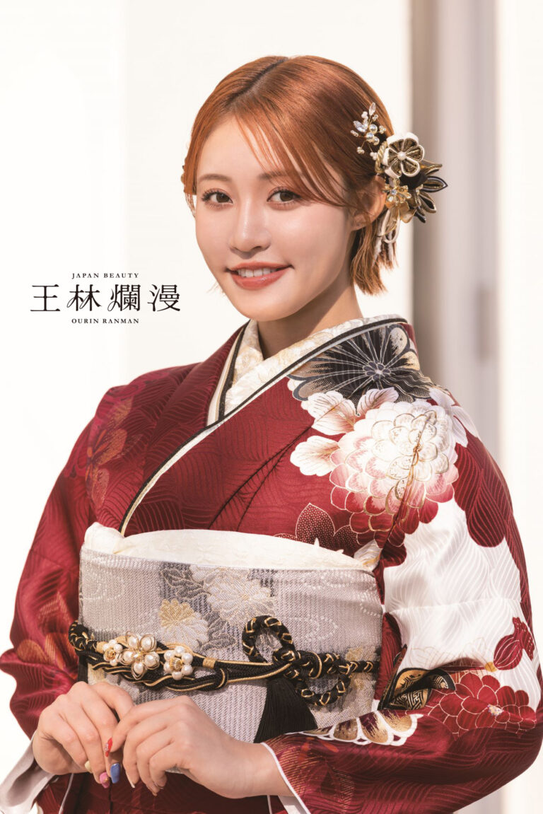 日本の伝統的な着物を着た人が微笑んでいます。着物はワインレッド色で、白と金の花柄が描かれています。短い栗色の髪には、花をあしらったエレガントなヘアピンが飾られています。横には「日本美人 桜林万歳」という文字が書かれています。背景は柔らかくぼかされています。.