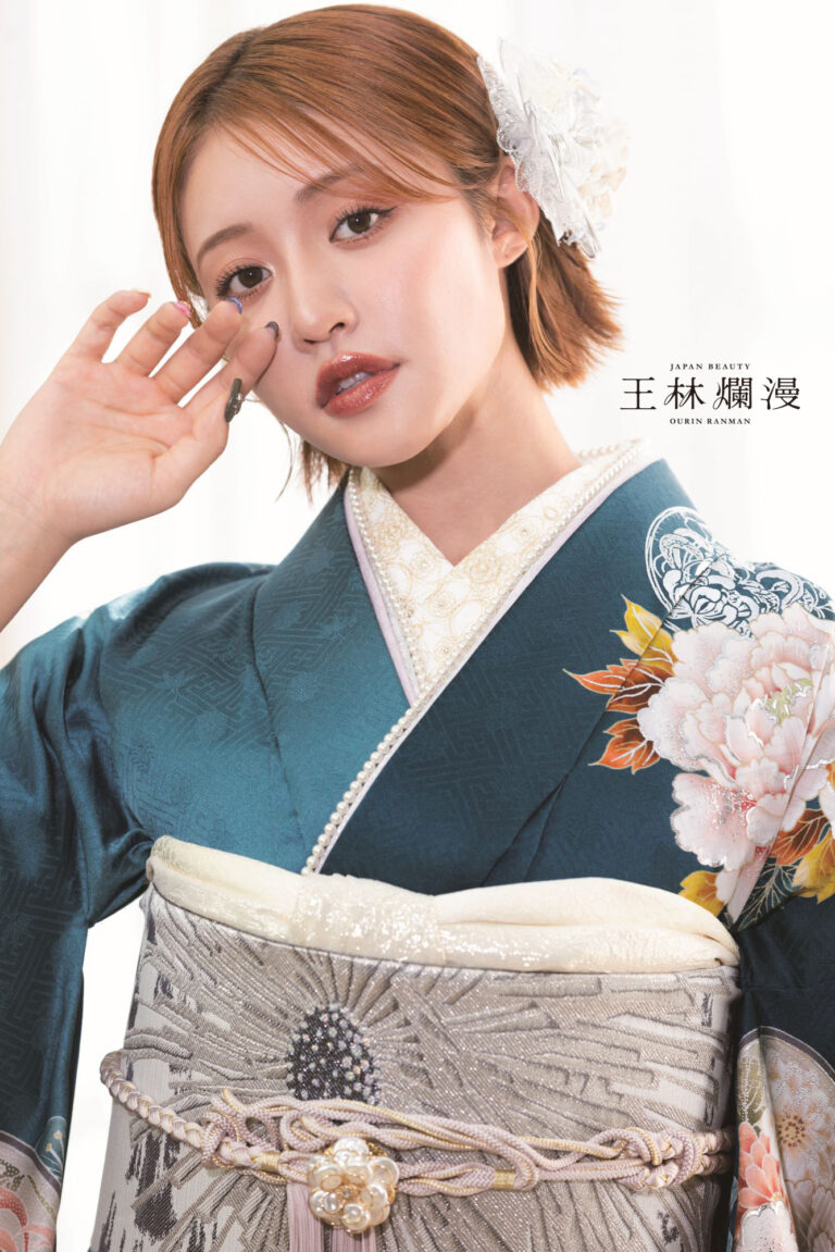 花の刺繍が施された青緑色の着物と濃い色の帯を締めた人物が、片手を顔の近くに持ってポーズをとっています。着物には白とピンクの色合いの大きくて複雑な花が描かれています。人物は短い髪に白い花のアクセサリーを飾っています。控えめなメイクと光沢のある唇をしています。.