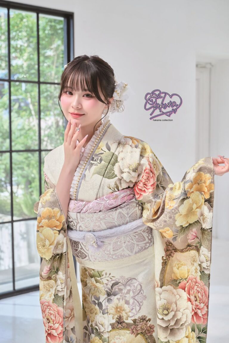 花柄の着物を着た女性が室内でポーズをとっています。着物には黄色、ピンク、オレンジの色合いの大きな色とりどりの花が描かれています。彼女は片手で顔を触り、白い花の髪飾りをつけています。背景には緑の景色が見える大きな窓があります。.