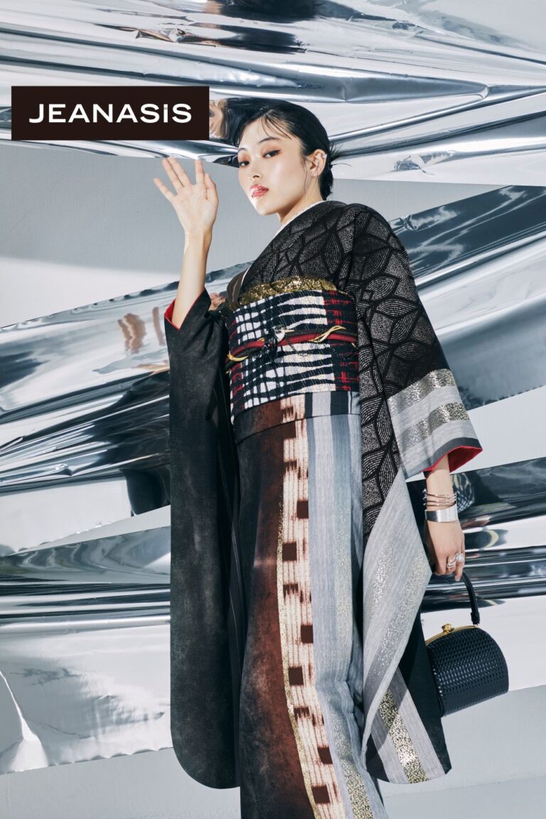 光沢のあるメタリックな背景を背景に、おしゃれな着物を着た人物がポーズをとっています。着物には複雑な模様と大胆な帯があしらわれています。人物は小さな黒いハンドバッグを持ち、メイクと髪型もスタイリッシュです。左上隅に「JEANASIS」という文字が表示されています。.