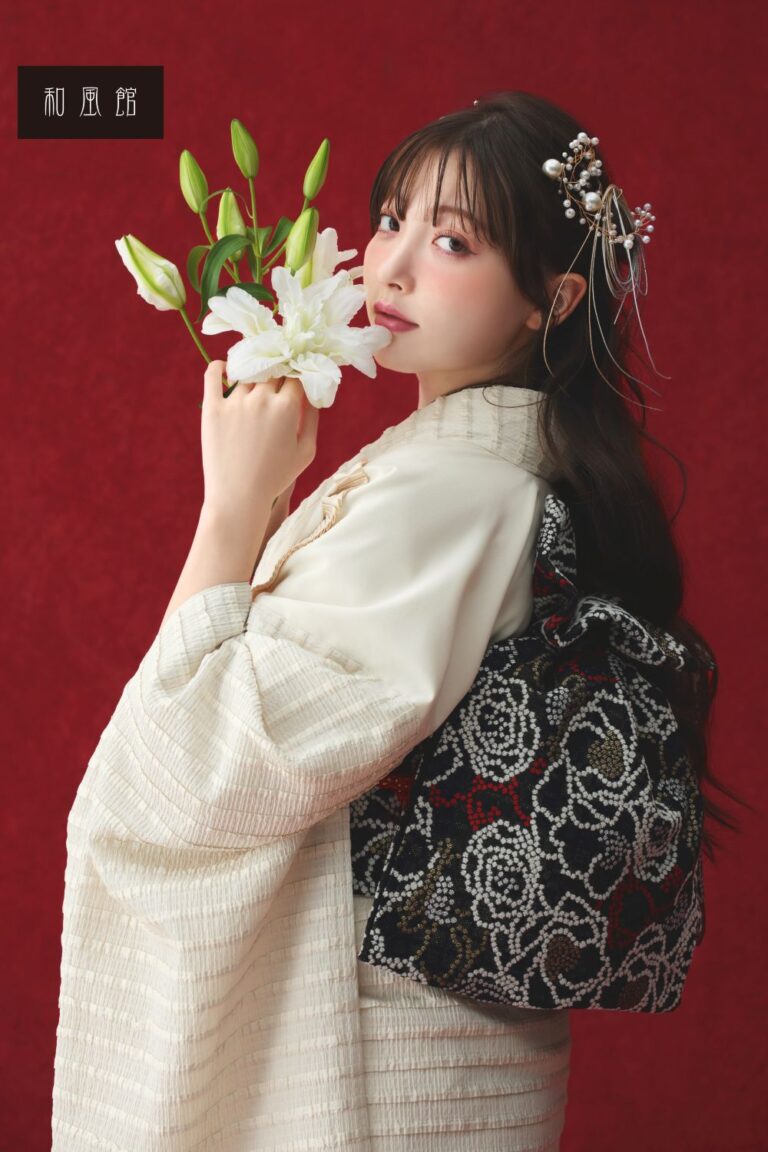 クリーム色の着物を着た女性が、鮮やかな赤の背景に白いユリの花束を持っている。彼女の黒髪には複雑な花のヘアピースが付いており、優雅にカメラを見つめている。彼女の背中には、25WF11 と識別される黒と白の模様のバッグが掛けられている。.