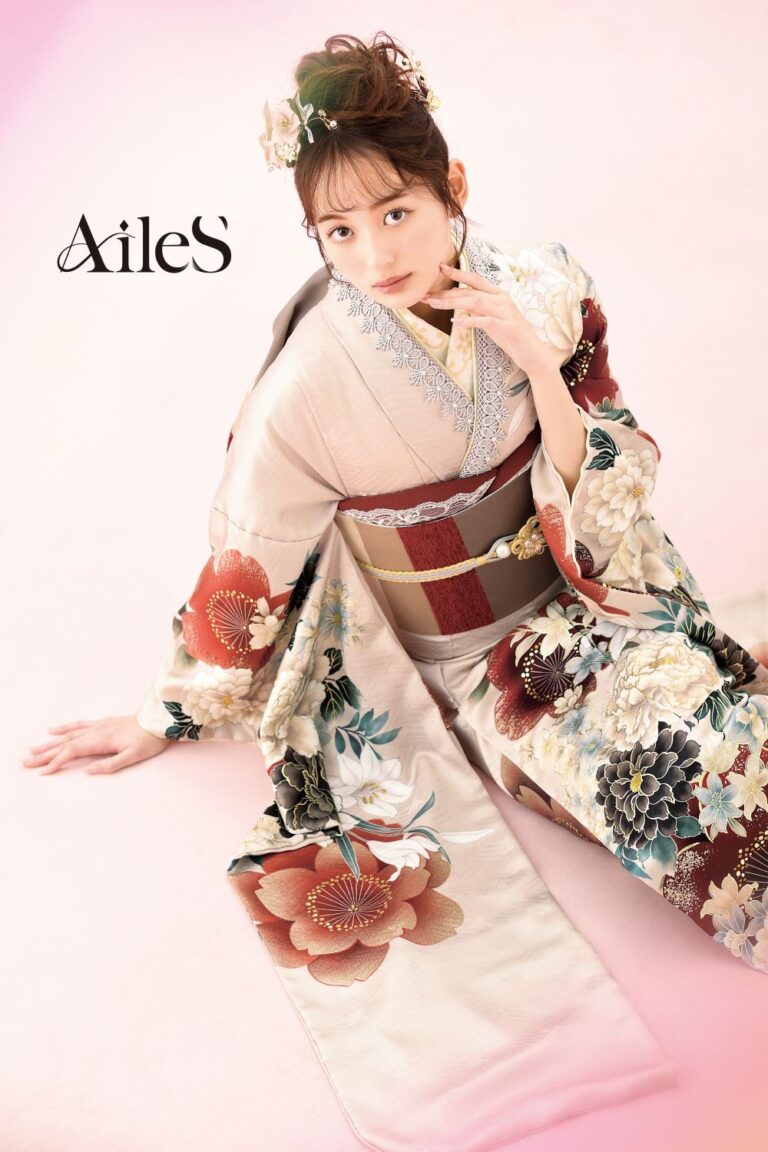 エレガントな花柄の着物を着た女性が淡いピンクの床に座っている。髪には花をあしらい、指をそっと唇に当てながら穏やかな表情でカメラを見つめている。上には筆記体で「AileS」と書かれている。着物には赤、青、クリーム色の複雑な花柄が描かれている。.