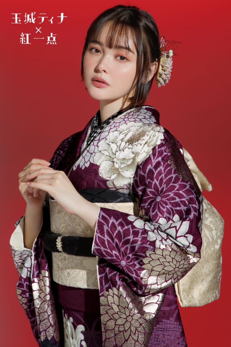 紫色の花柄の着物を着た女性が赤い背景に立っています。彼女の髪には花飾りが飾られています。彼女は真剣な表情をしており、両手は優雅に前に出ています。左上に日本語の文字が見えます。.