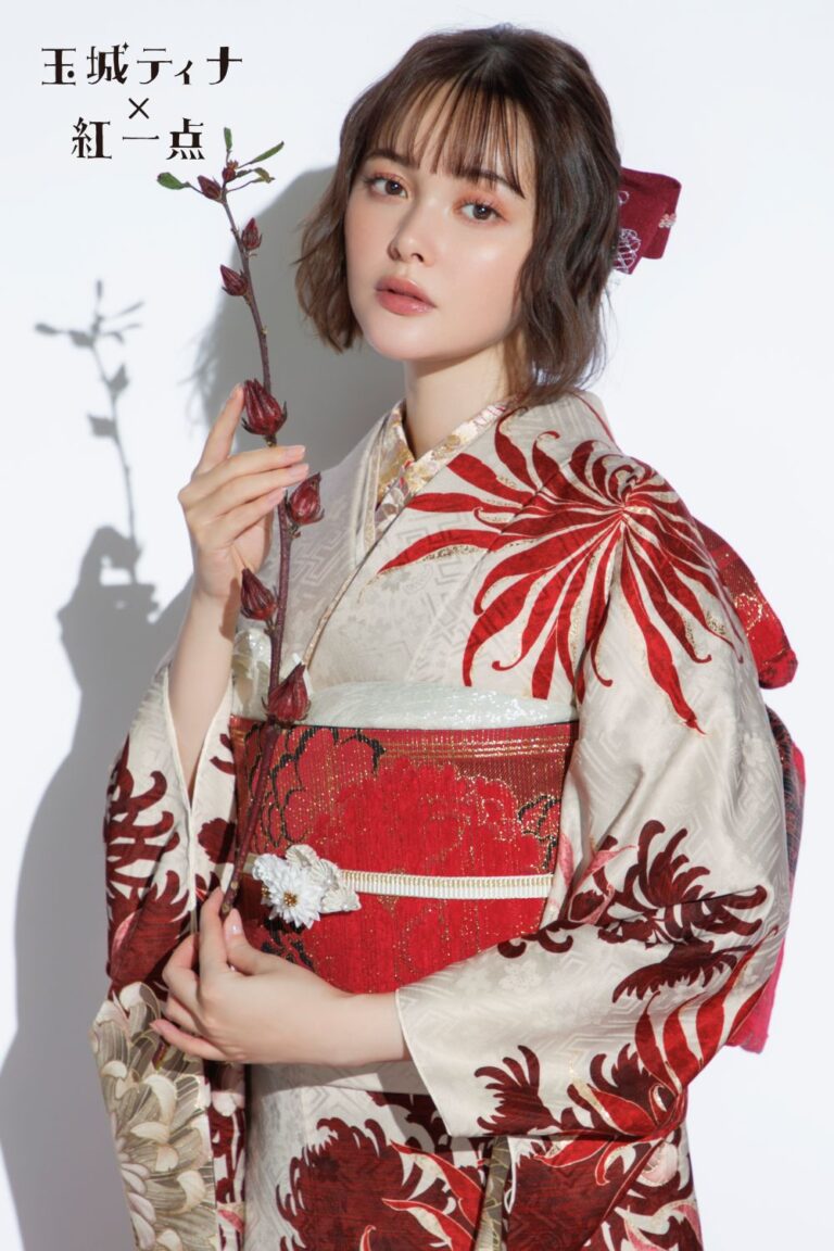 赤と白の花柄の伝統的な着物を着て、葉の付いた枝を持っている人物。着物には複雑な赤い模様があり、赤い帯を締めています。人物は短い髪に赤いリボンをつけています。日本語のテキストは左側にあります。背景は無地の白です。.