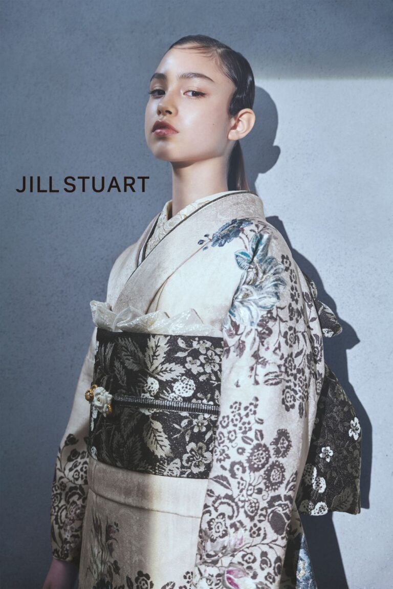 淡いブルーの背景に、上品な花柄の伝統衣装と濃い帯を身に着けた若者が立っています。髪は後ろになでつけ、自信に満ちた表情で前を見つめています。画像の左側には、黒い文字で「JILL STUART」と縦書きされています。.