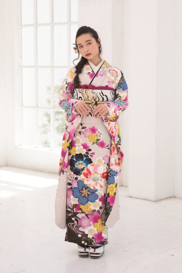 明るい部屋に、ピンク、青、黄色の花柄があしらわれた色鮮やかな着物を着た女性が立っている。長い黒髪をヘアアクセサリーで少し束ね、少し横を向いている。背景には白い壁と大きな窓が見える。.