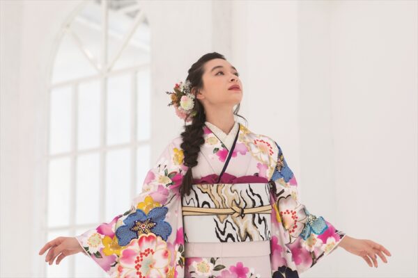 色鮮やかな花柄の着物を着た女性が、両腕を軽く広げて立っている。長い髪は花飾りで編まれている。彼女は、大きなアーチ型の窓を背景にした明るい白い部屋の中で、上を見上げている。.