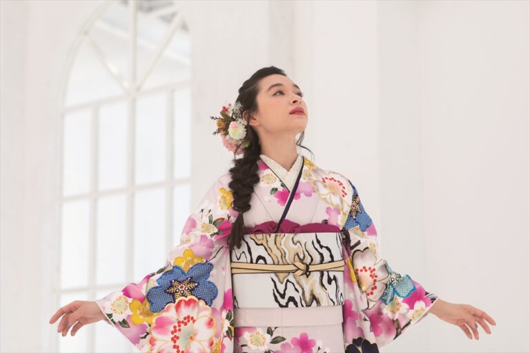 色鮮やかな花柄の着物を着た女性が、両腕を軽く広げて立っている。長い髪は花飾りで編まれている。彼女は、大きなアーチ型の窓を背景にした明るい白い部屋の中で、上を見上げている。.