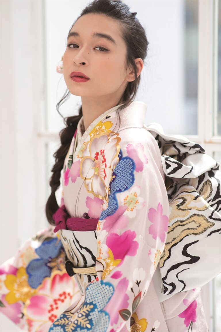 長い黒髪を三つ編みにした女性が、ピンク、黄色、青のカラフルな花柄があしらわれた白い着物と、白黒の渦巻き模様が入った大きな帯を締めている。室内で、柔らかな化粧を施し、自信に満ちた表情でカメラを見つめている。.