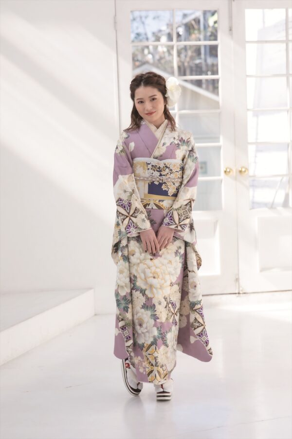 若い女性が、日差しが差し込む室内に立っている。パステルカラーの紫と白の花柄の着物に、柄物の帯を締めている。髪には花を挿し、優しく微笑んでいる。伝統衣装の下には白いスニーカーを履いている。背景は大きな窓から明るく照らされている。.