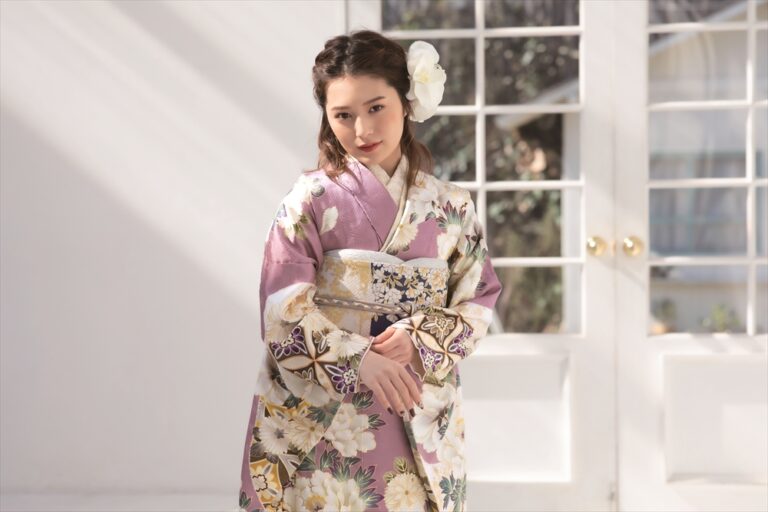 柔らかなウェーブのかかった黒髪に大きな白い花をあしらった若い女性が、淡い紫の花柄の着物をまとっている。彼女は室内で、陽光が差し込むガラス張りのドアの前に立ち、両手を優しく組んで、少し横を向いている。.