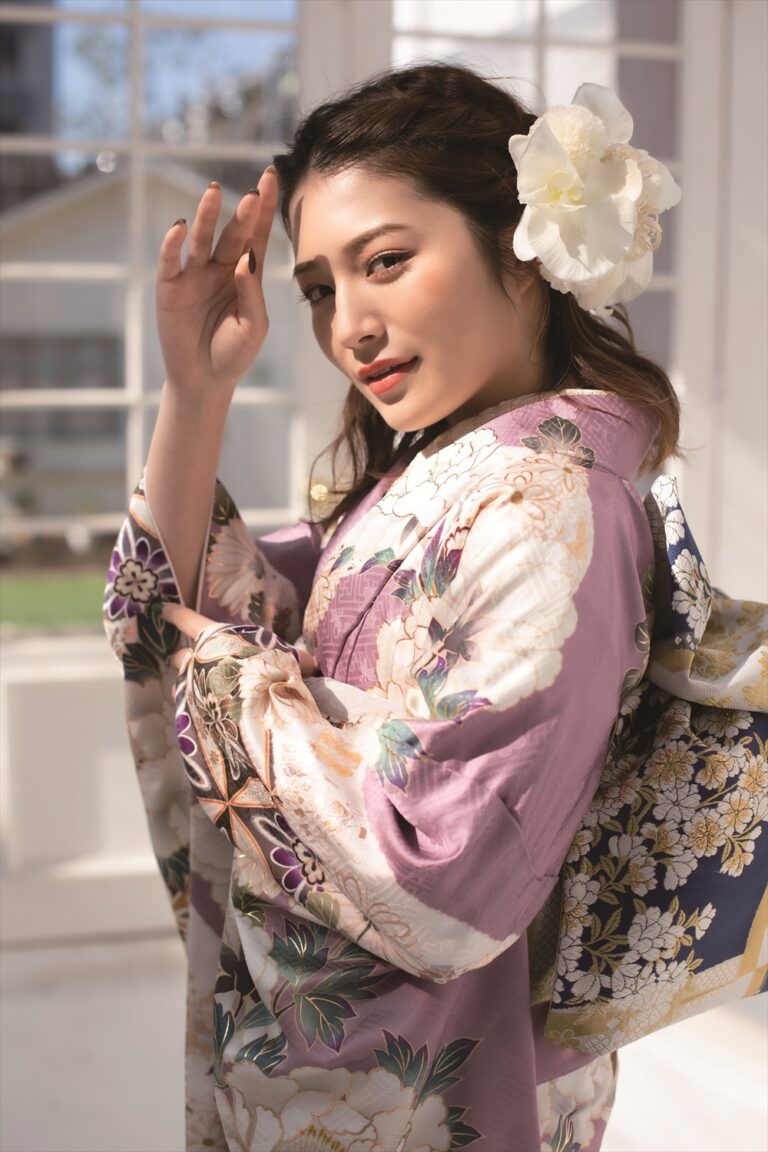 薄紫の花柄の着物を着た若い女性が室内でポーズをとっている。長い黒髪に大きな白い花のアクセサリーを飾り、頭のそばで手を上げている。背後の高い窓から差し込む陽光が、柔らかく優雅な雰囲気を醸し出している。.