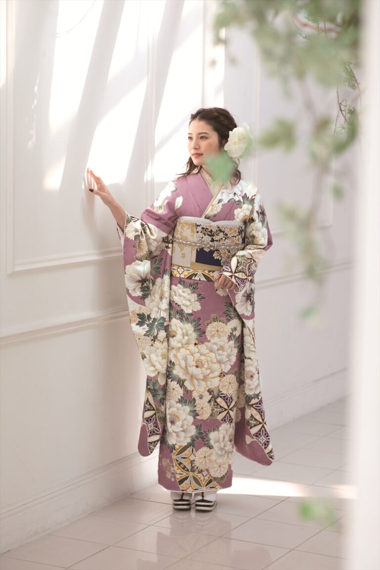 薄紫色の花柄の着物に金帯を締めた女性が、陽光に照らされた白い壁のそばに立ち、軽く壁に触れている。花の髪飾りをし、白い草履を履いている。柔らかな光と緑が、穏やかで優雅な雰囲気を醸し出している。.