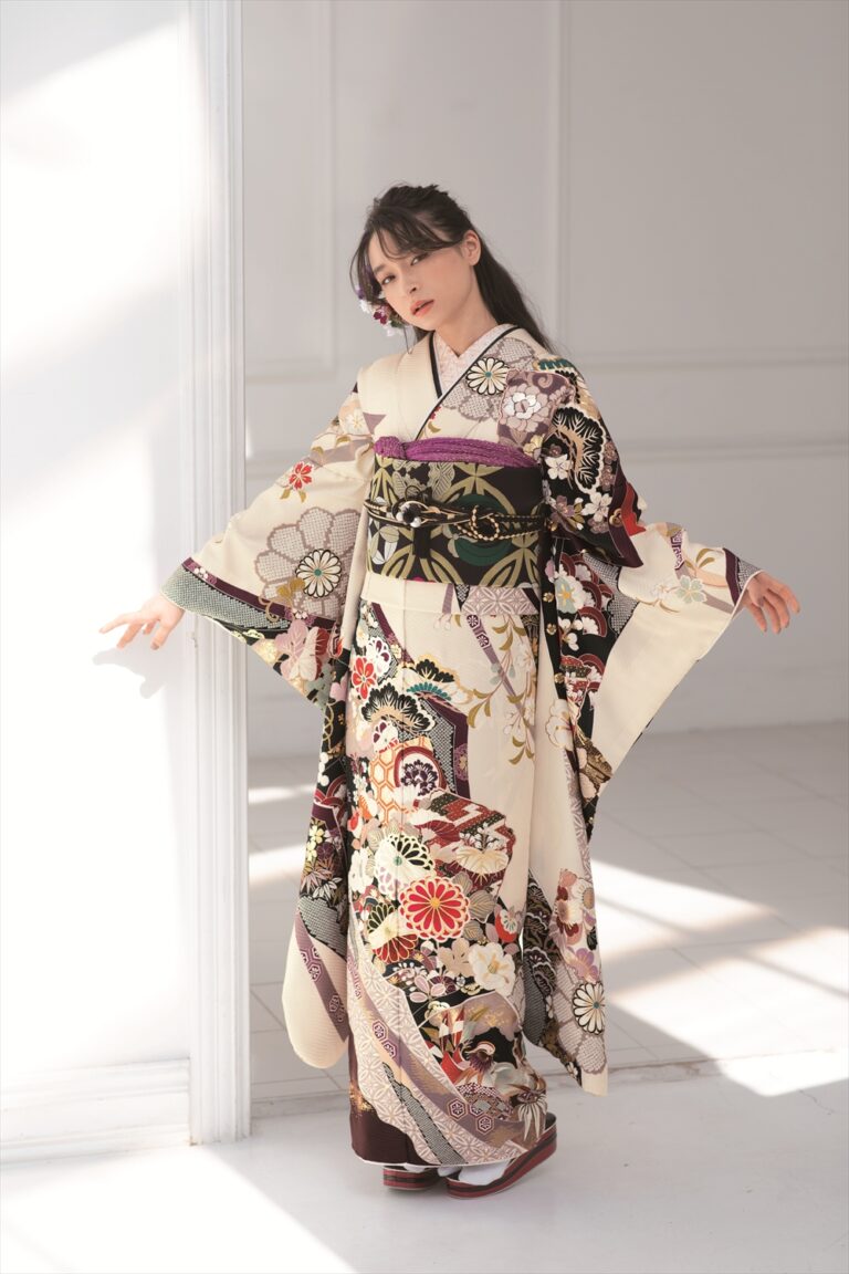 明るい日差しが差し込む部屋の中で、クリーム色の着物を着た女性が立っている。色鮮やかな花柄や幾何学模様があしらわれた着物を着て、落ち着いた表情で横を向いている。黒髪を少し後ろに束ね、両腕を優しく伸ばして着物の袖を覗かせている。.