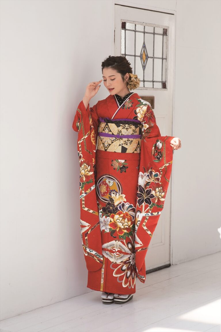 室内に、金、白、黒の花柄があしらわれた鮮やかな赤い着物を着た女性が立っている。髪には飾りが付けられ、優しく顔を撫でながらうつむいている。背景には白い壁と、ステンドグラスの窓が映る扉が描かれている。.