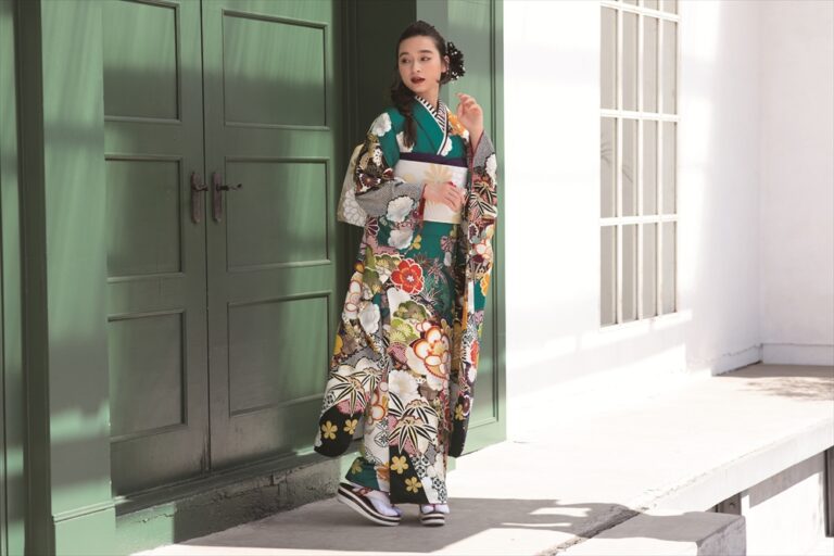 陽光を浴びる緑の両開きの扉のそばに、色鮮やかな花柄の着物に帯を締め、草履を履いた女性が立っている。髪は花飾りできれいにまとめられ、穏やかで上品な表情をしている。.