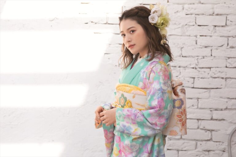 大きな花飾りをつけた色鮮やかな花柄の着物をまとった若い女性が、白いレンガの壁を背に立っている。彼女は帯の脇で両手を合わせ、優しくカメラを見つめている。差し込む陽光が、風景を明るく照らしている。.