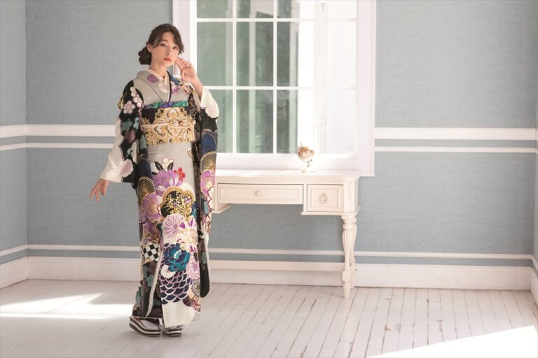 色鮮やかで華やかな花柄や幾何学模様の着物を着た女性が、白い机と大きな窓の近くでポーズをとっています。淡いブルーの部屋に白い縁取りが施され、柔らかな光が差し込み、優雅で落ち着いた雰囲気を醸し出しています。.
