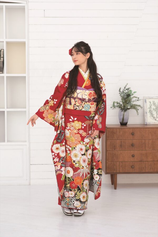 鮮やかな赤い花柄の着物を着た若い女性が、室内に立って左を向き、片手で指さしている。長い黒髪に赤い花が飾られている。彼女の後ろには、観葉植物が置かれた木製のドレッサーと、棚のある白い壁がある。.