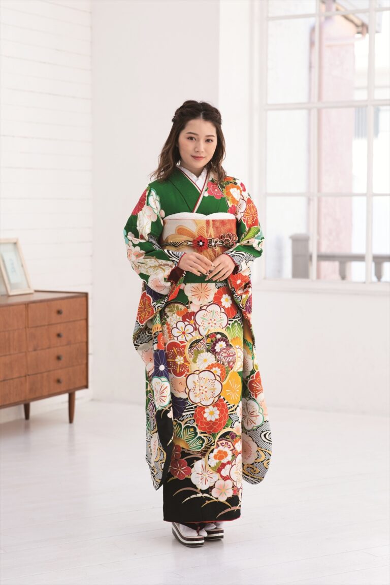 緑、赤、黄、白の模様が入った色鮮やかな花柄の着物を着た茶色の長い髪の女性が、室内に立っている。彼女は両手を前に組んでカメラを見つめている。部屋は白い壁と大きな窓、そして木製の戸棚が備わっている。.