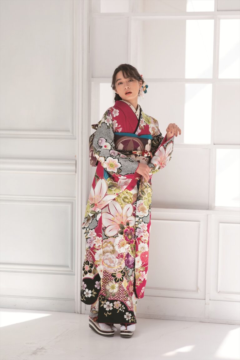 白い羽目板の壁のそばに、若い女性が立っている。赤、ピンク、黒、クリーム色の鮮やかな花柄の着物を着て、髪には花飾りが付けられている。片腕を上げ、もう片方の腕を腰に当て、背後から差し込む光の中、彼女は優しく前方を見つめている。.