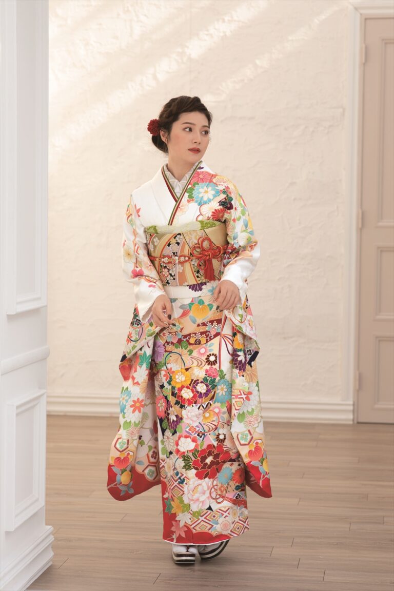 白い部屋の中で、色鮮やかな花柄の着物に金の帯を締めた女性がいる。赤い髪飾りをつけた凝った髪型で、左を向いている。部屋の雰囲気は明るく、木の床と明るい色の壁とドアが印象的だ。.