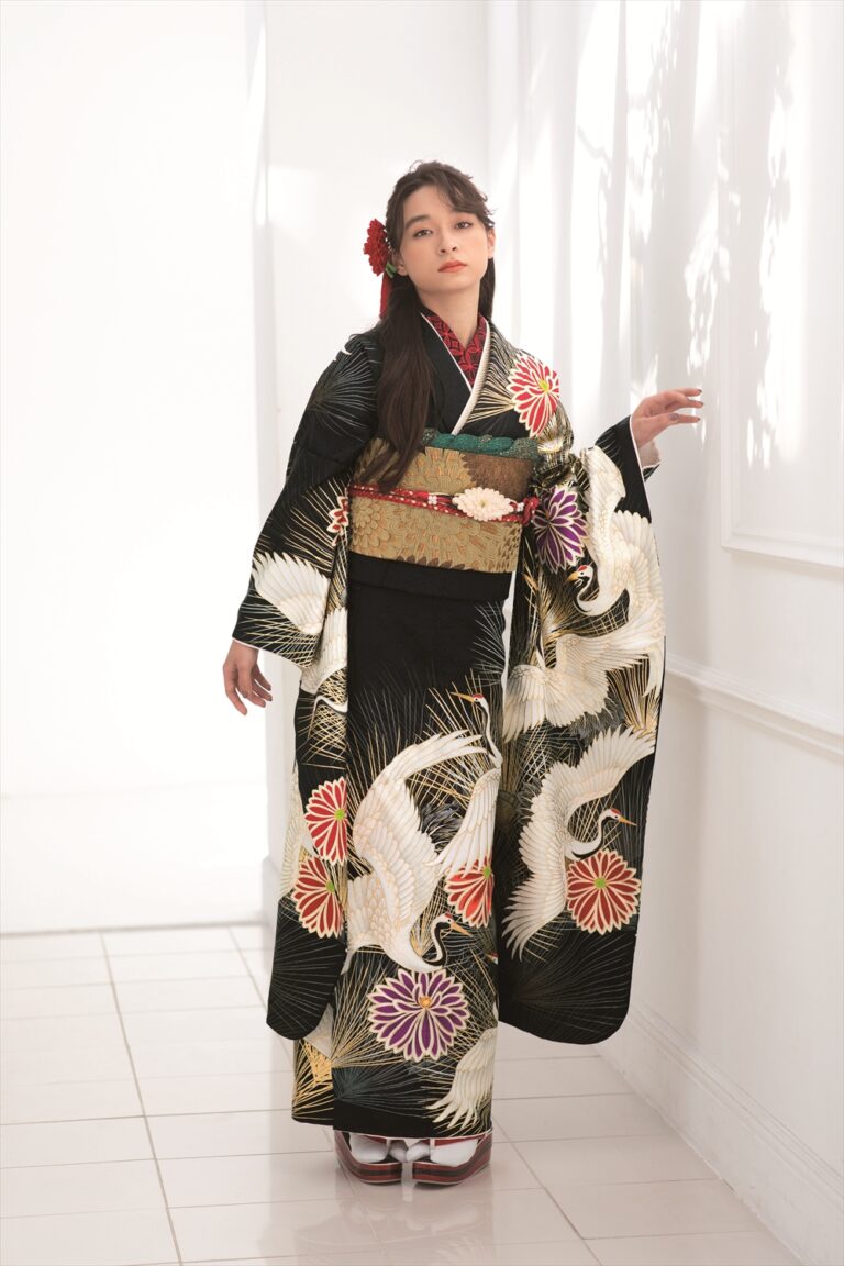 白い鶴と色とりどりの花が飾られた伝統的な黒い着物を着た女性が、室内に立っている。長い黒髪に赤い花の髪飾り、白い足袋に木靴を履いている。窓から差し込む光が、壁に柔らかな影を落としている。.