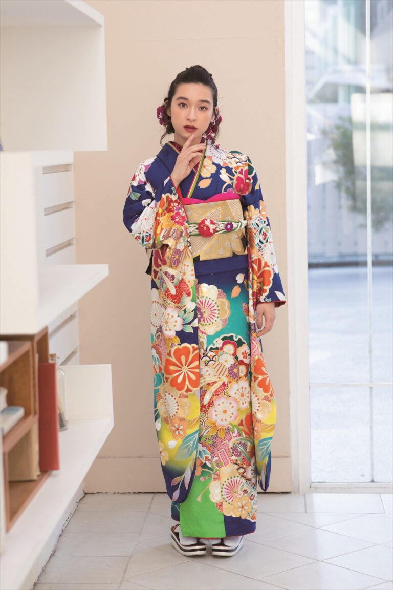 色鮮やかな花柄の着物に金の帯を締めた女性が室内に立っている。片手を顔の近くに置き、カメラを見つめている。髪には花飾りが飾られている。左側には棚と本が並び、背後の窓から陽光が差し込んでいる。.