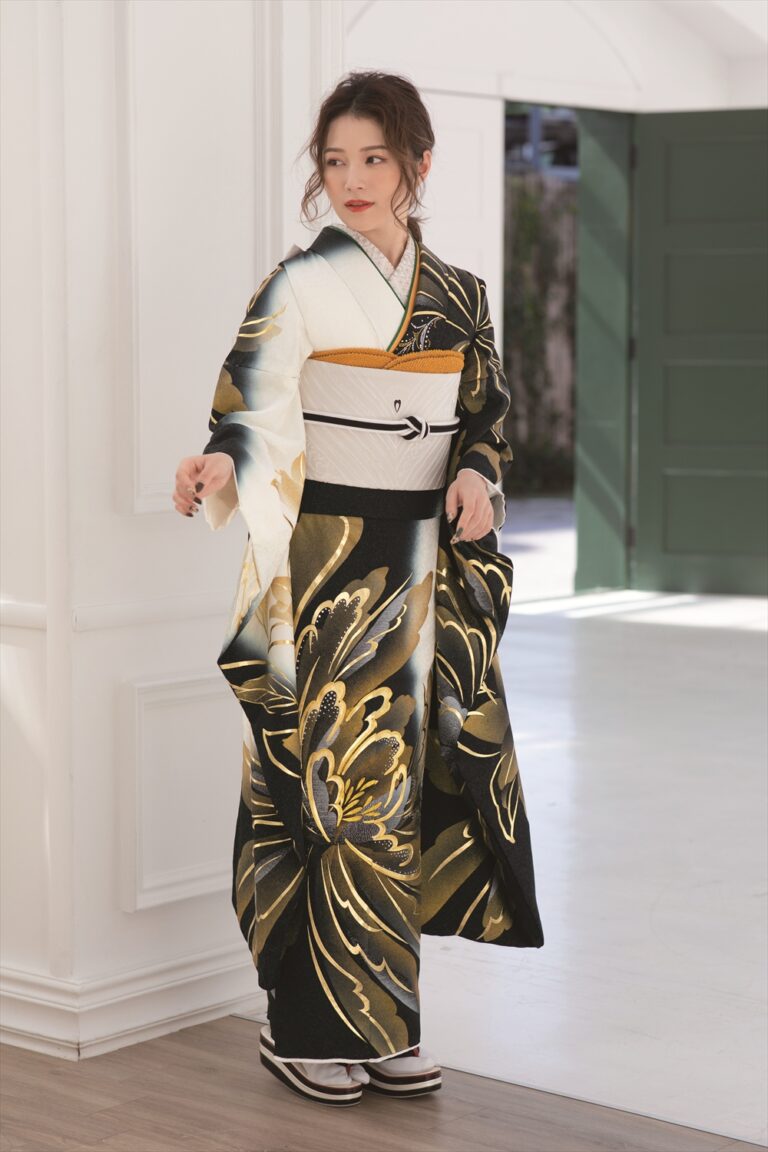 黒と金の花柄の着物に白と金の帯を締めた女性が室内に立っている。ウェーブのかかった茶色の髪をアップにし、伝統的な白い足袋と黒い草履を履いている。部屋は明るく、白い壁と、背景には開いた緑色のドアがある。.