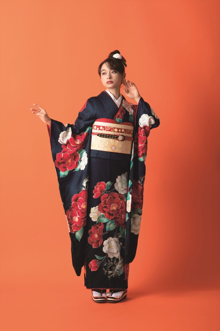 鮮やかな赤と白の花柄があしらわれた紺色の着物を着た女性が、オレンジ色の背景に立っています。腰には華やかな帯を締め、白い足袋と伝統的な履物を履いています。髪はアップスタイルにまとめられ、白い髪飾りが飾られています。.