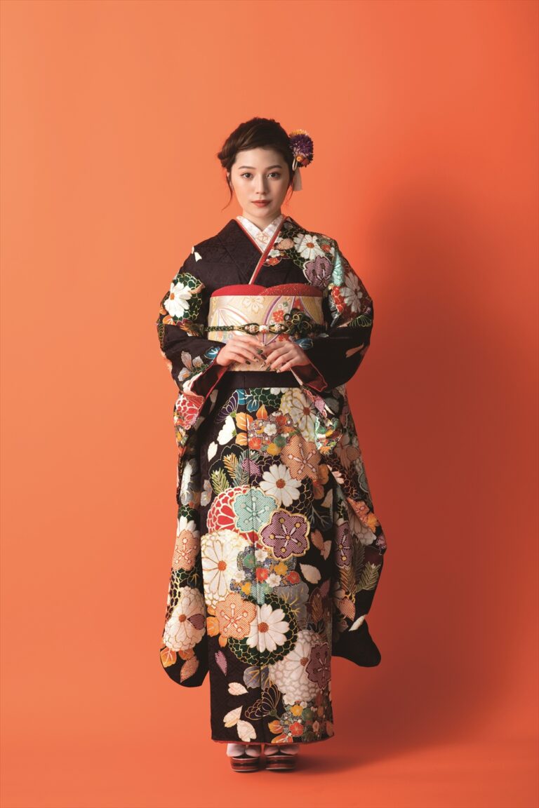 オレンジ色の背景に、色鮮やかで華やかな花柄の着物を着た女性が立っています。髪は紫色の花飾りできちんとまとめられ、両手を優しく前に組んだ穏やかな表情をしています。草履を履いています。.