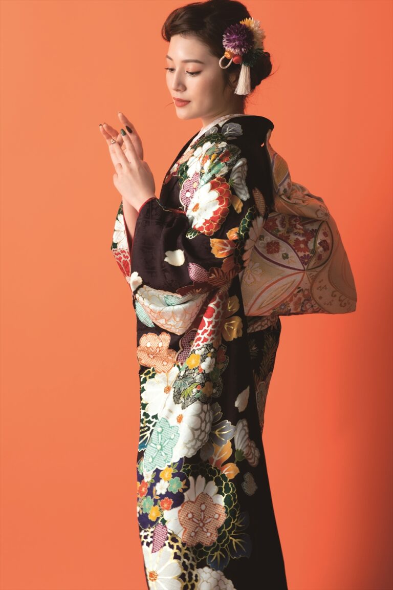 オレンジ色の背景に、色鮮やかな花柄の着物に大きくて華やかな帯を締めた女性が横顔で立っている。髪はきちんとセットされ、花や飾りのヘアピースで飾られている。彼女は両手を見つめ、穏やかで優雅な表情を浮かべている。.