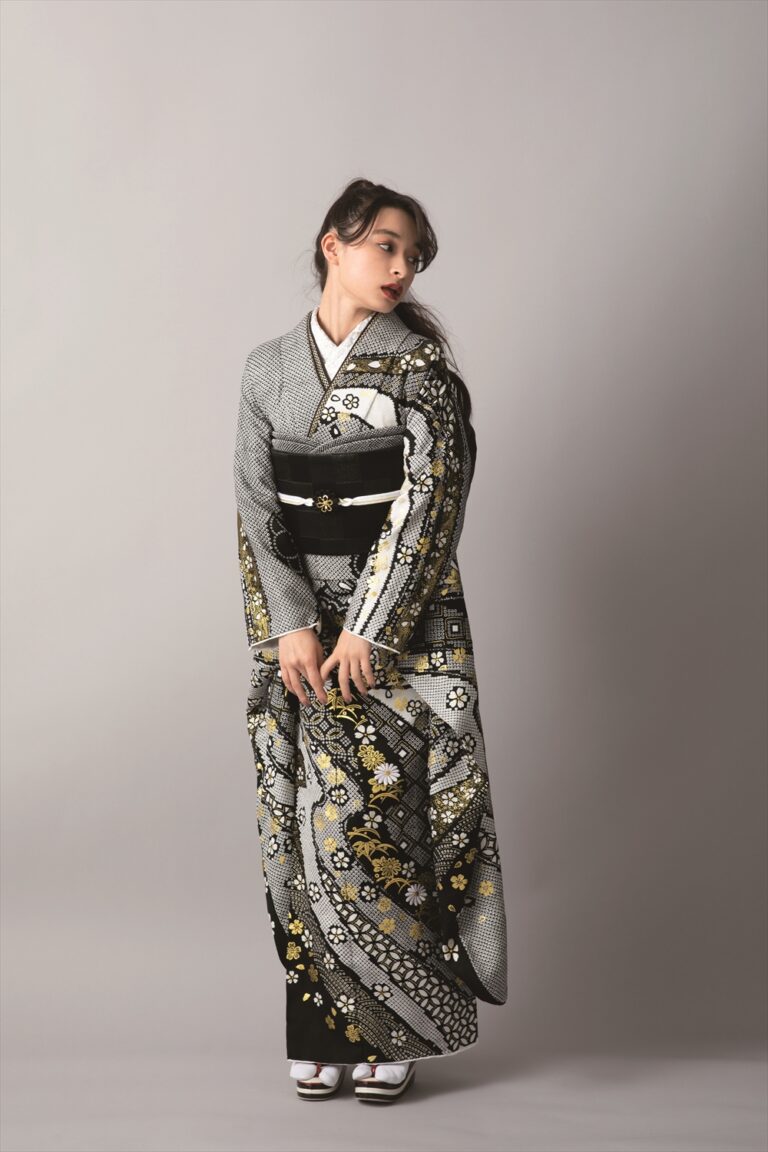 複雑な花柄と幾何学模様があしらわれた黒、白、金の正装着物を着た女性が、無地の背景に立ち、右を向いている。両手を前に出し、白い足袋と草履を履いている。.