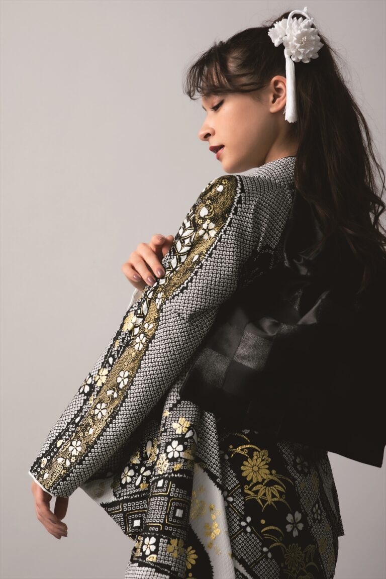 横顔の女性は、黒、白、金の花柄の着物に黒帯を締め、長い黒髪に白い花の髪飾りを添えている。彼女は明るい背景に、目を閉じて優雅なポーズをとり、優雅さと落ち着きを漂わせている。.