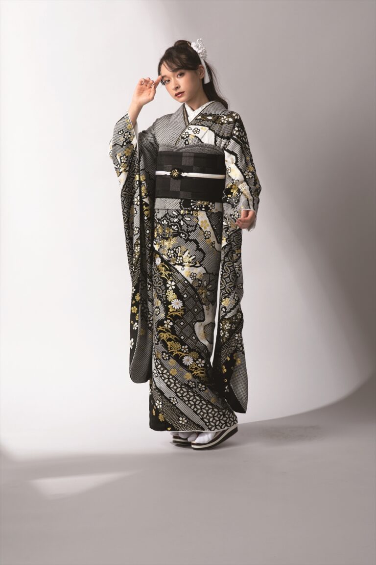 黒、白、金の花柄があしらわれた正装の着物を着た女性が、シンプルな背景に立っています。白い髪飾り、白い足袋、そして伝統的な草履を身につけ、片手を頭の横に、もう片方の手を腰に当てています。.