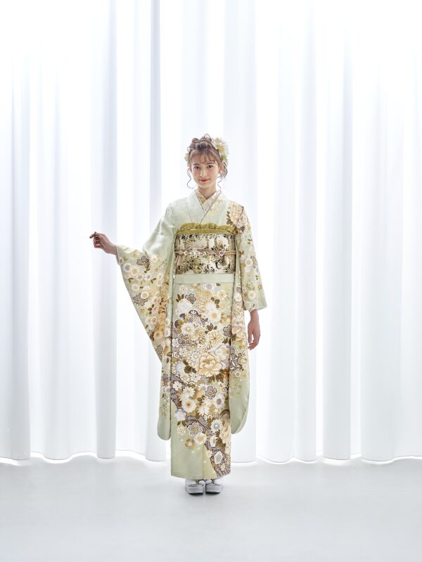 白いカーテンの前に、金、白、茶色の花柄があしらわれた淡い緑色の着物を着た女性が立っている。髪には飾りの簪が飾られ、白い草履に白い足袋を履いている。.