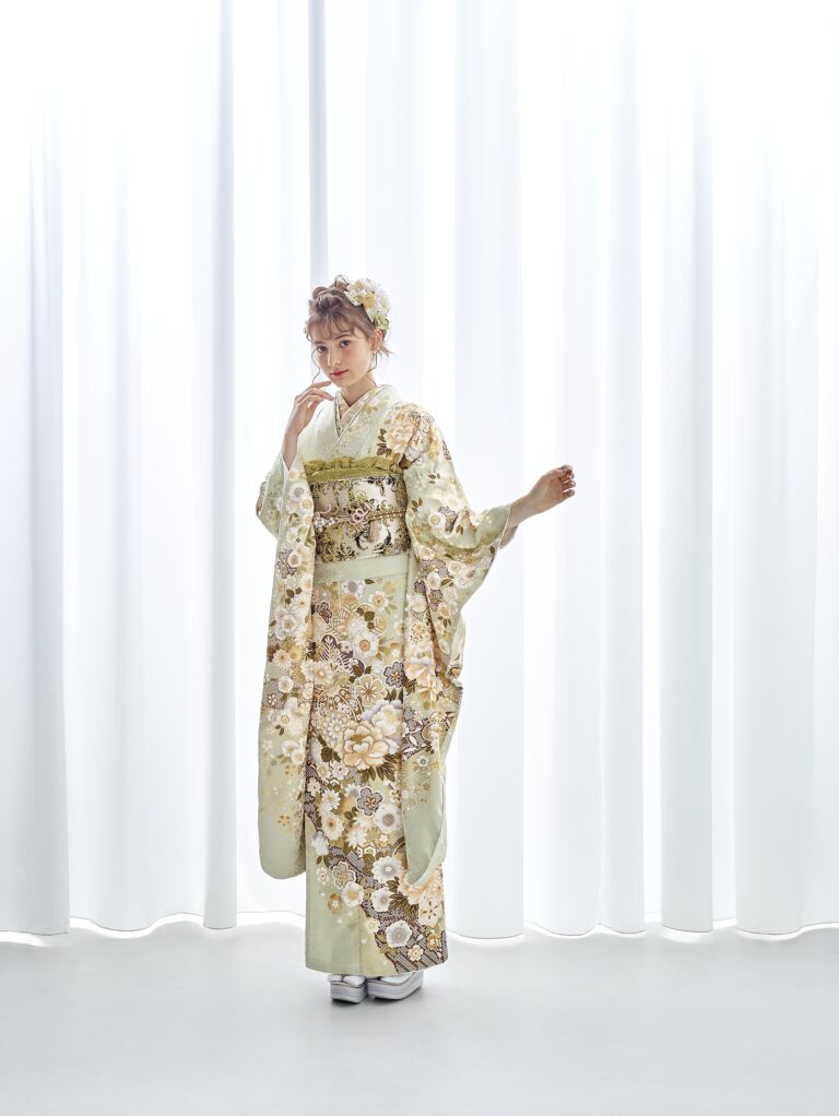 白いカーテンの前に、金と白の花柄があしらわれた淡い緑色の着物を着た女性が立っている。薄茶色の髪に花飾りをし、白い草履を履いている。片手を顔に近づけ、もう片方の手を少し上げてポーズをとっている。.