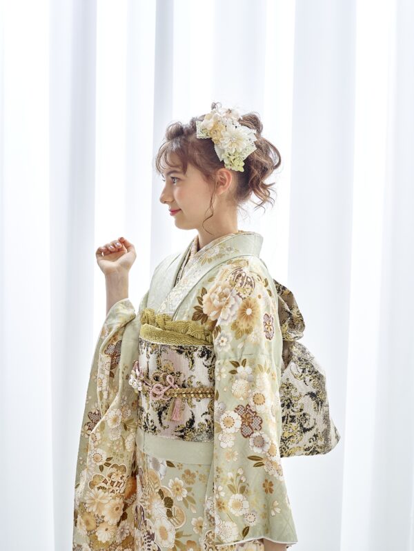 若い女性が横顔で立っています。薄緑の花柄の着物に、幅広の帯と後ろに飾りリボンを結んでいます。髪には白とパステルカラーの花を添えてアップにしています。柔らかな光が差し込む白いカーテンを背景に、ポーズをとっています。.