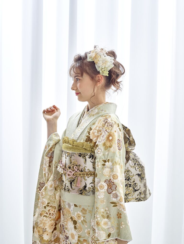 若い女性が横顔で立っています。薄緑の花柄の着物に、幅広の帯と後ろに飾りリボンを結んでいます。髪には白とパステルカラーの花を添えてアップにしています。柔らかな光が差し込む白いカーテンを背景に、ポーズをとっています。.