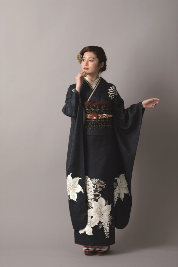大きな白い花柄があしらわれた黒い着物を着た女性が、ニュートラルな背景を背に立っている。髪は飾りの簪できちんとセットされ、片手を挙げて着物の長い袖を際立たせながら、横を向いて優雅なポーズをとっている。.