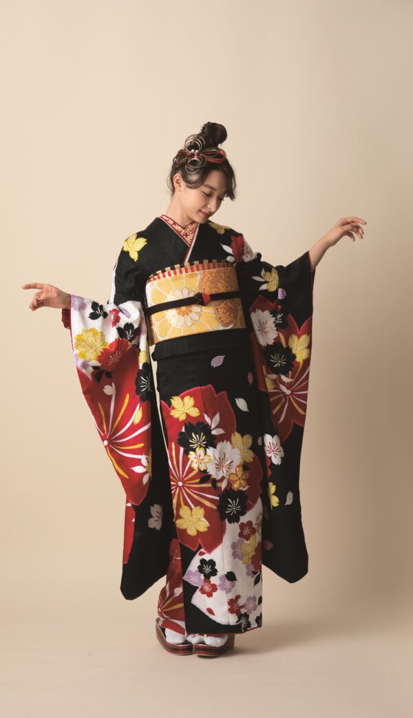 ベージュの背景に、白、黄、赤の花柄があしらわれた黒の着物（商品番号AI22-22）を着た若い女性がポーズをとっています。伝統的な髪飾り、華やかな帯、下駄を身につけ、着物の袖を披露しています。.