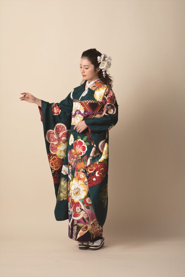 鮮やかな花柄があしらわれた濃い緑色の着物を着た女性が、無地のベージュの背景に立っています。白い花の髪飾りをし、髪はきちんとセットされています。彼女はやや下を向き、左を向いて優雅なポーズをとっています。.