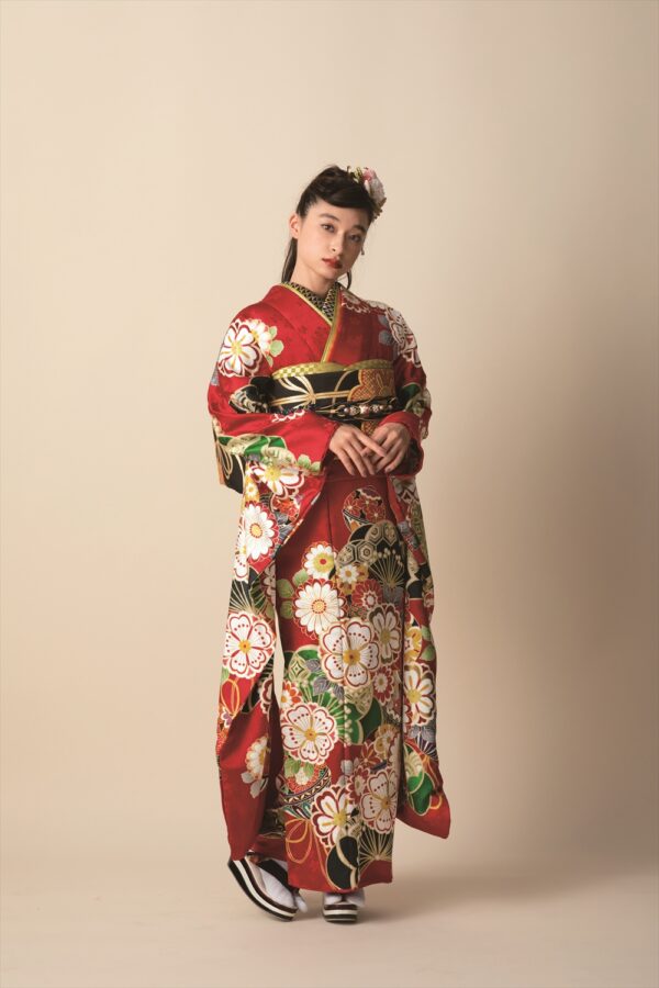 ベージュの無地の背景に、鮮やかな赤い着物を着た若い女性が立っています。着物には、色とりどりの大きな花柄があしらわれています。黒と金の帯、白い足袋、黒い草履を履いています。髪は花の髪飾りできれいにまとめられています。.