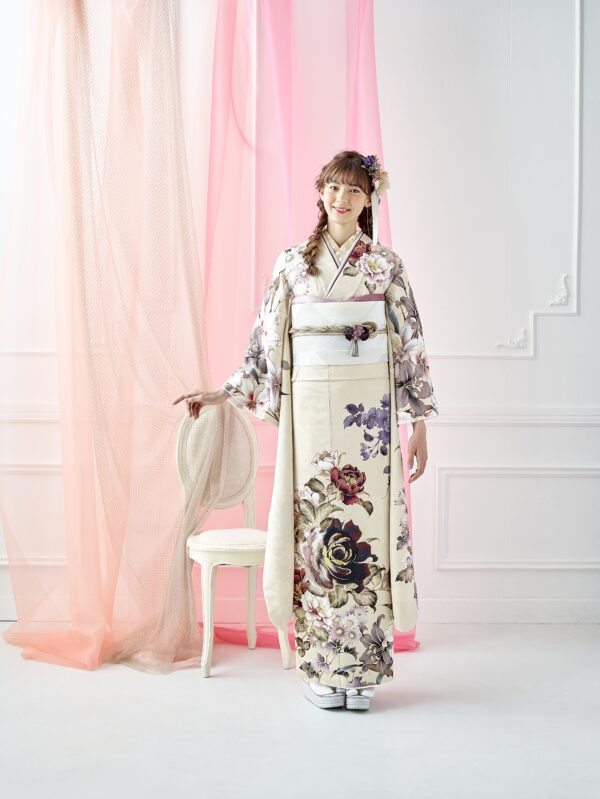 クリーム色の大きな花柄の着物を着た女性が室内に立っている。髪には花と三つ編みが飾られ、白い椅子と透け感のあるピンクのカーテンが白い装飾の壁にかかっている。明るく優雅な光景だ。.